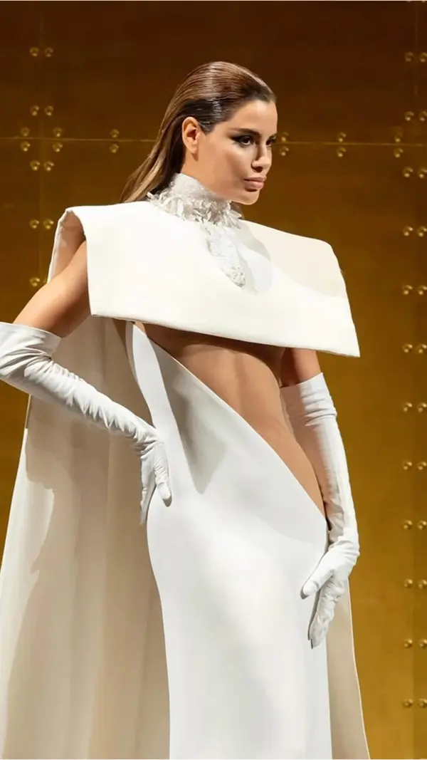 femme portant une longue robe blanche et une longue cape. Découpe asymétrique sur son ventre.